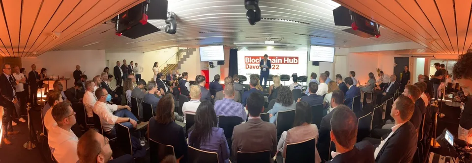 Panoramic photo of the same main room during the Forum, packed with a standing-room audience facing the stage